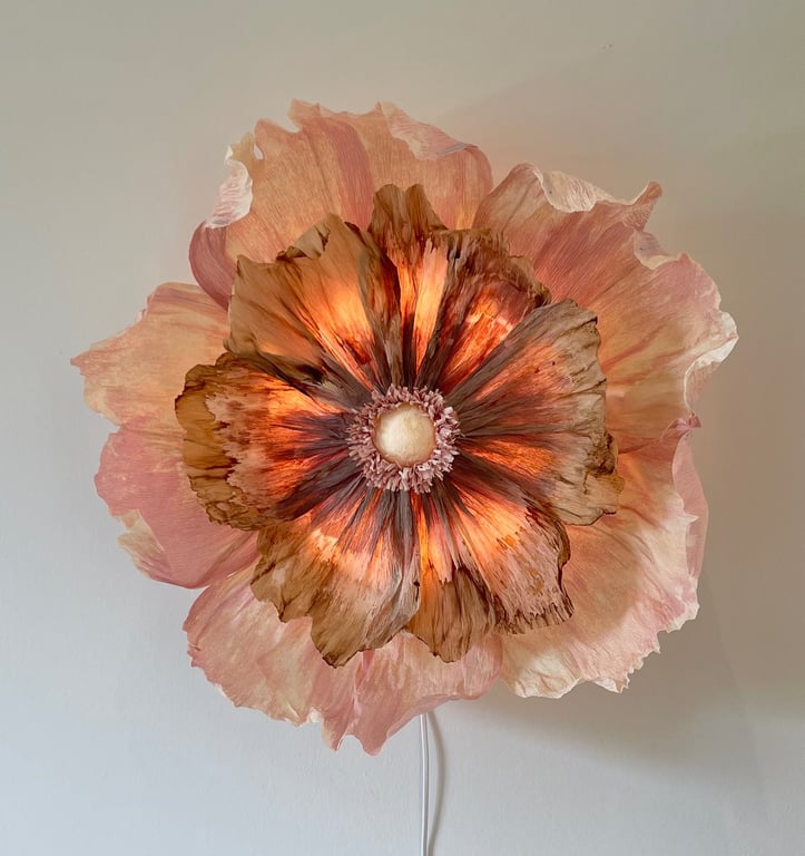 Lysende lampe i form af en stor, realistisk blomst med rosa og brunlige kronblade mod en hvid væg.