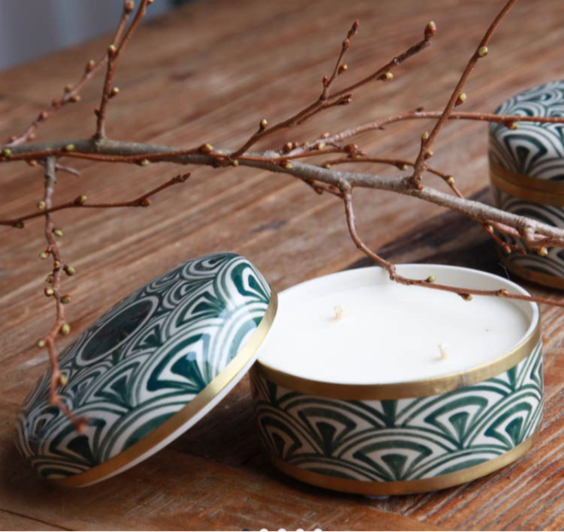 To-lugede duftlys i dekorative, grønne og hvide mønstrede metalbeholdere med guldkanter, placeret på et træbord med en kviste ovenpå. (2)
