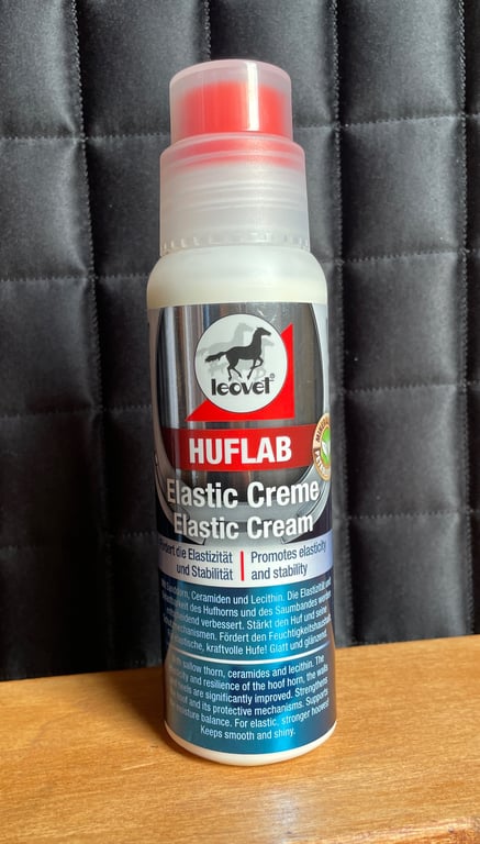 Flaske med Leovet Huflab Elastic Creme til hove med tekst på dansk og engelsk, placeret på træbord foran sort polstret baggrund.