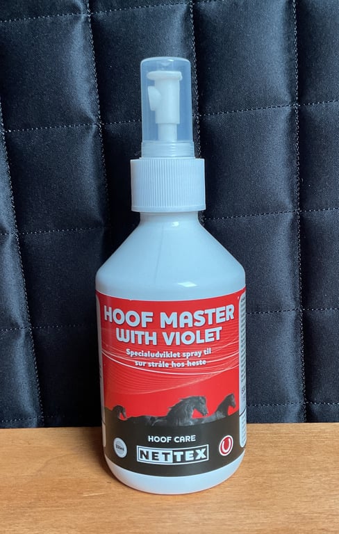 Flaske med HOOF MASTER WITH VIOLET hoveplejespray til heste med rød og hvid label, placeret på et træbord foran en sort quiltet baggrund.