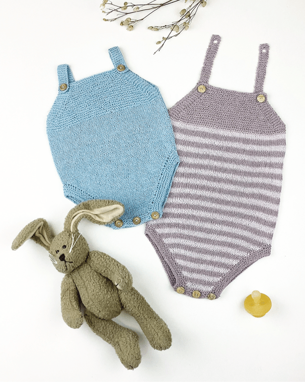 To strikkede babybodys i blåt og lilla med knapper, en grå bamse-kanin og en pacifier på en hvid baggrund med tørrede blomster.