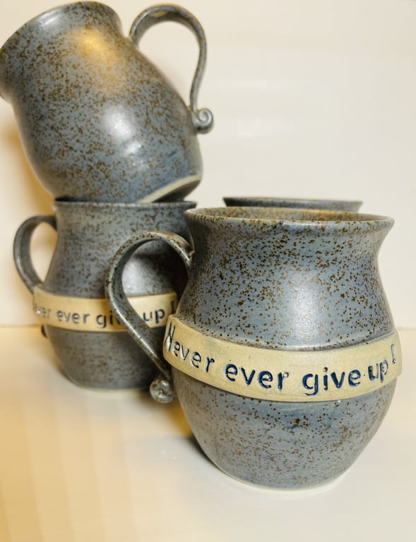 Gråmelerede keramikkrus med hank, hvorpå teksten "Never ever give up!" er indgraveret på en beige stribe rundt om krusene.