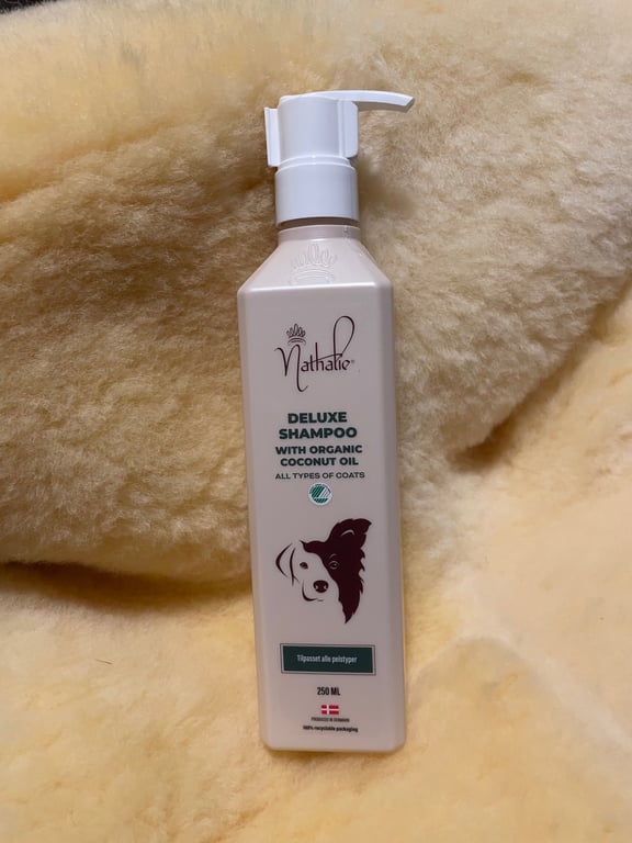 Flaske med Nathalie Deluxe Shampoo med økologisk kokosolie til alle pels typer, 250 ml, placeret på et lyst, blødt uld- eller pelslignende underlag.