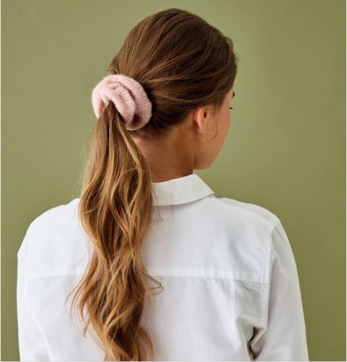 Kvinde set bagfra med langt, lyst bølget hår samlet i en hestehale med en lyserød hårscrunchie, iført en hvid skjorte mod en grøn baggrund. (2)