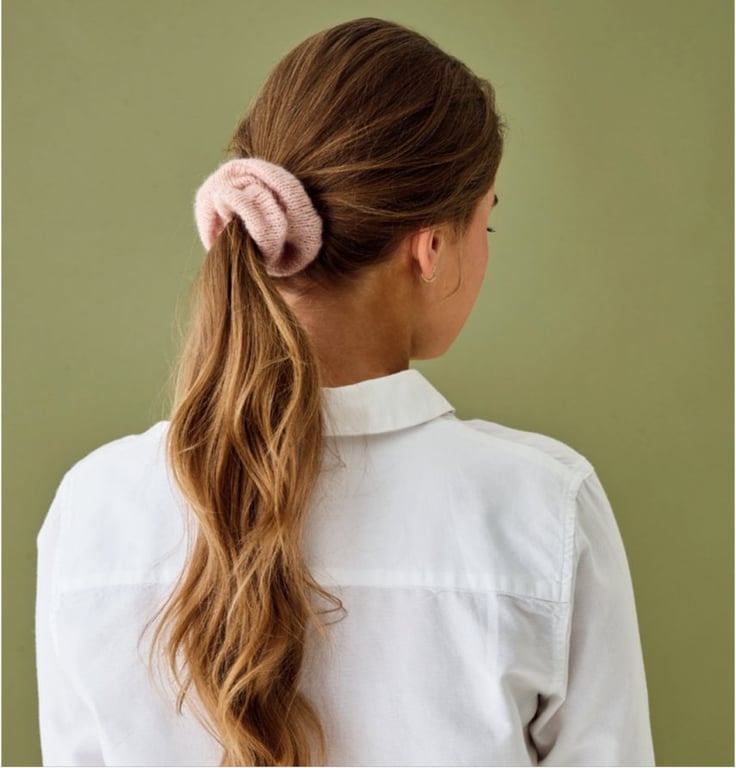 Kvinde set bagfra med langt, lyst bølget hår samlet i en hestehale med en lyserød hårscrunchie, iført en hvid skjorte mod en grøn baggrund.