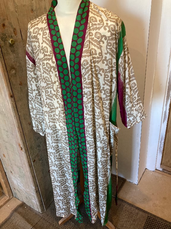 Lang, åben kimono med beige og hvide blomsteragtige mønstre, grønne og lilla detaljer langs kanterne og på ærmerne, vist på en hvid mannequin i et rum med træmøbler. (2)