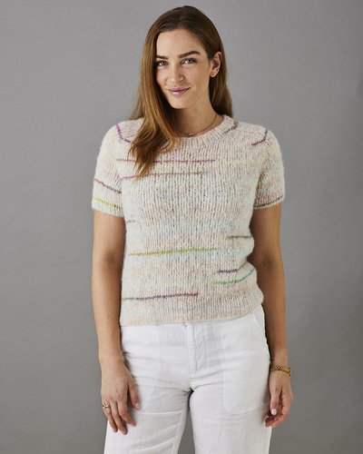 Kvinde med langt brunt hår iført kortærmet, strikket sweater med pastelfarver og hvide bukser, stående foran grå baggrund.