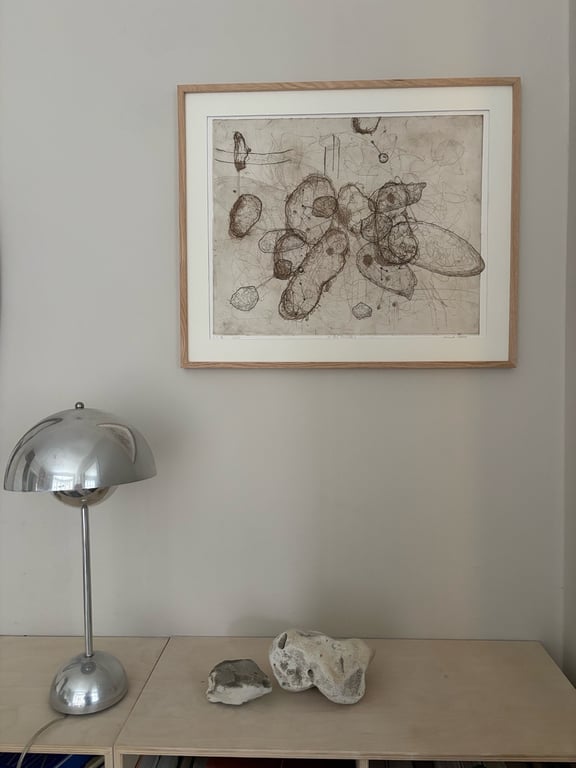 Væg med abstrakt tegning i lyst træramme, moderne sølvfarvet bordlampe og to dekorative sten på lyst bord.