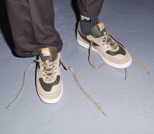 Nærbillede af et par sneakers med beige og grønne detaljer, uagtede skosnørebånd og mørkebrune bukser på gråt gulv. (1)