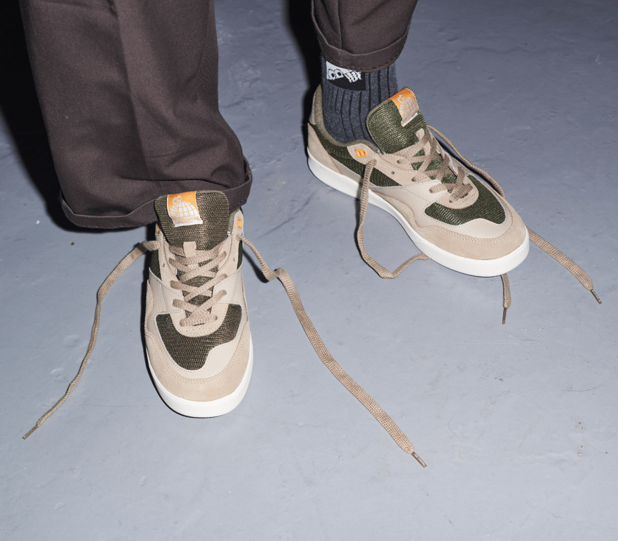 Nærbillede af et par sneakers med beige og grønne detaljer, uagtede skosnørebånd og mørkebrune bukser på gråt gulv.