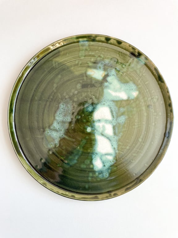 Rund keramisk tallerken med grøn glasur og et mønster af lyseblå og hvide pletter på overfladen, fotograferet ovenfra mod en hvid baggrund.