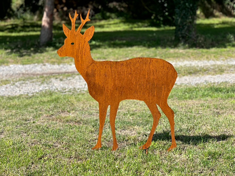 Rustfarvet metalfigur af en hjort med gevir, placeret på en græsplæne med træer i baggrunden.