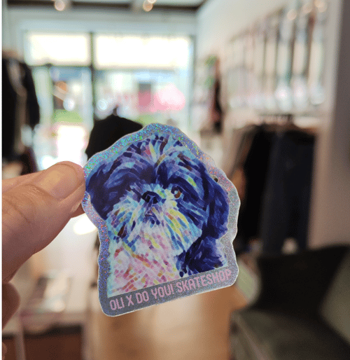 Hånd der holder et glitrende klistermærke med et malet billede af en hund og teksten "OLI X DO YOU! SKATESHOP" i et lyst, sløret butiksmiljø. (1)
