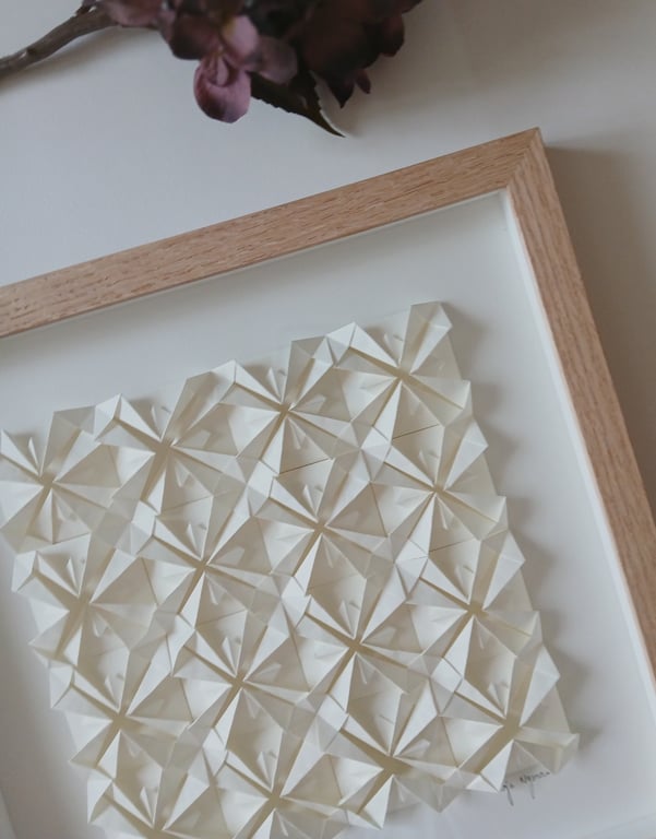 Billedramme med tredimensionelt, mønstret papirkunst bestående af foldede hvide firkanter.