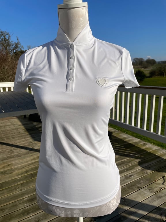 Hvid kortærmet poloshirt med fire knapper og lille logo på brystet, vist på en mannequin udendørs på en træterrasse i solskin.