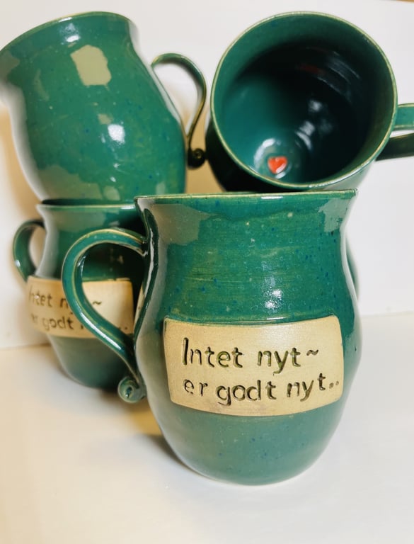 Grønne keramikkrus med beige mærker, hvorpå der står "Intet nyt er godt nyt." Et krus viser et lille rødt hjerte indvendigt.