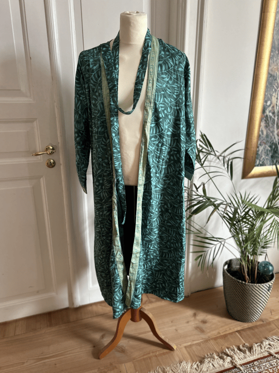 Grøn kimono med lyst mønster hængende på en mannequin i et lyst rum med trægulv, planter og hvid dør i baggrunden. (2)
