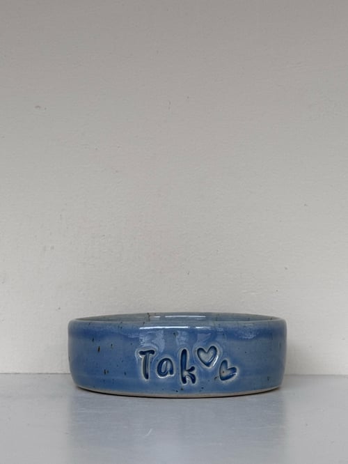 Blå keramisk skål med indgraveret tekst "Tak" og to hjerter, placeret på en hvid overflade mod en lys væg. (1)