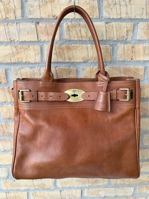 Brun lædertaske med to håndtag, dekorativ spænde og nøgleskede foran, hængende på en krog mod en murstensvæg.