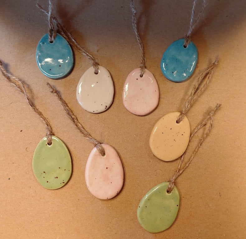 Billede af otte små, glaserede keramikvedhæng i forskellige pastel farver med groft jutesnor på neutral baggrund.