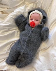 Baby iført en grå, strikket heldragt med hætte og luffer, liggende på et lyst lagen med en rød sut i munden. (1)
