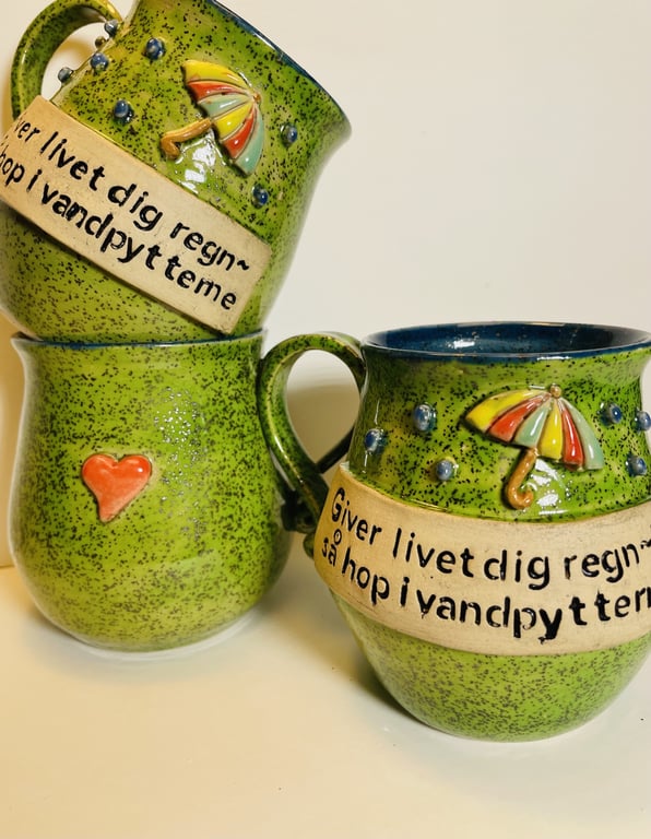 Tre grønne keramikkrus med tekst på dansk og dekorationer af paraplyer og et rødt hjerte.