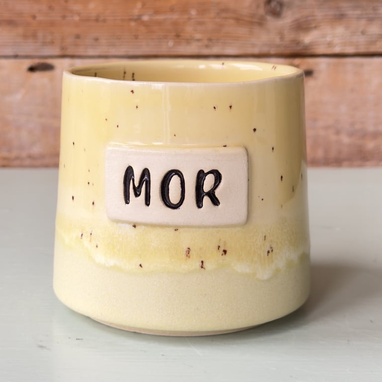 Gul keramikkrukke med teksten "MOR" på en rektangulær plade, stående på en lys overflade med træbaggrund.