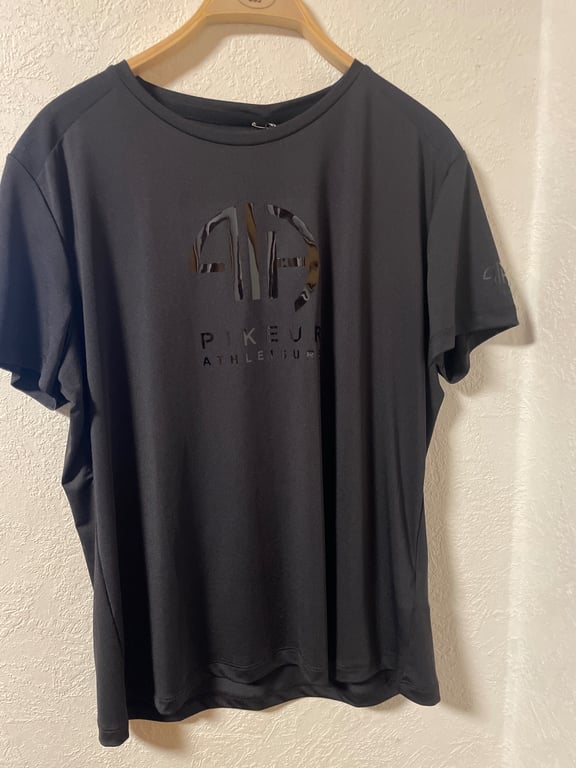 Sort t-shirt med rund hals og tryk af logo og tekst "Pikeur Athleisure" hængende på en bøjle mod en hvid væg.