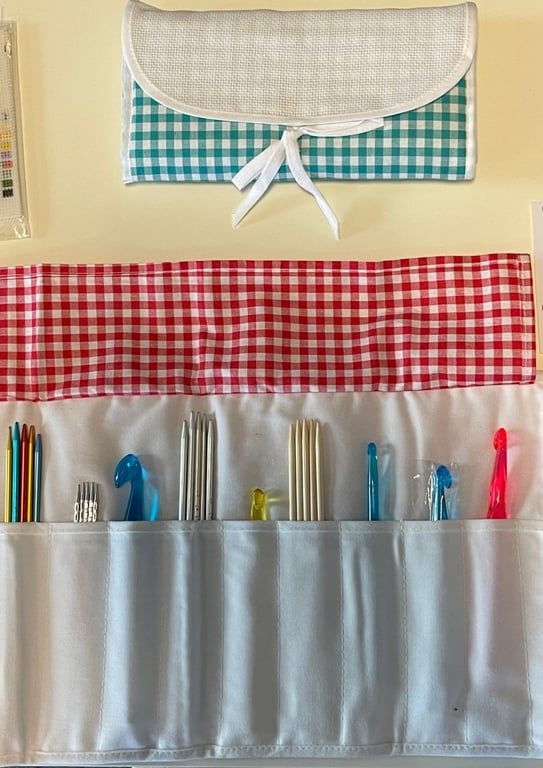 Strikkepinde og hæklenåle i forskellige størrelser organiseret i en stofrulle med rødt og hvidt ternet lommetryk, samt en sammenfoldet opbevaringspose øverst med blå og hvidt tern.