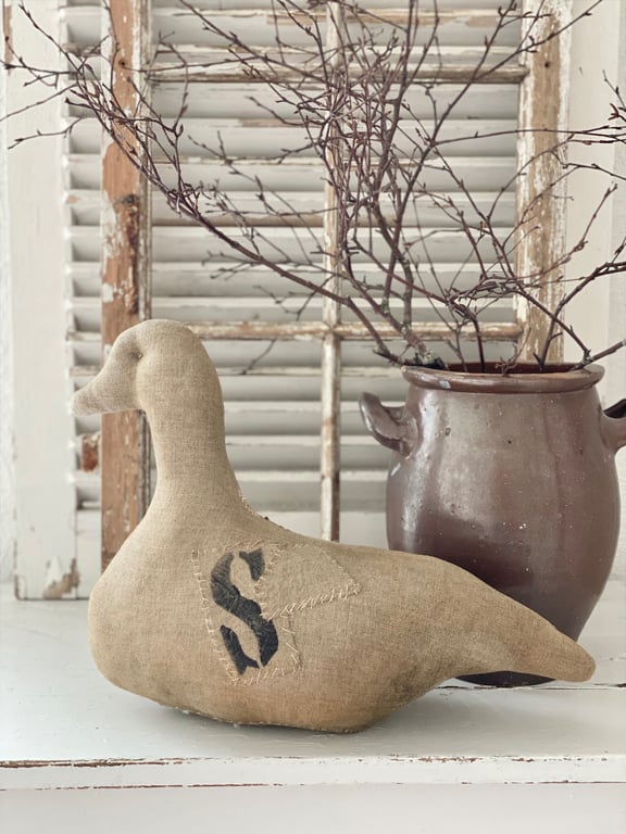 Håndlavet stofand med applikation af bogstavet "S" foran placeret på et hvidt bord foran en rustik krukke med tørre grene og et gammelt, afskallet vinduespanel.