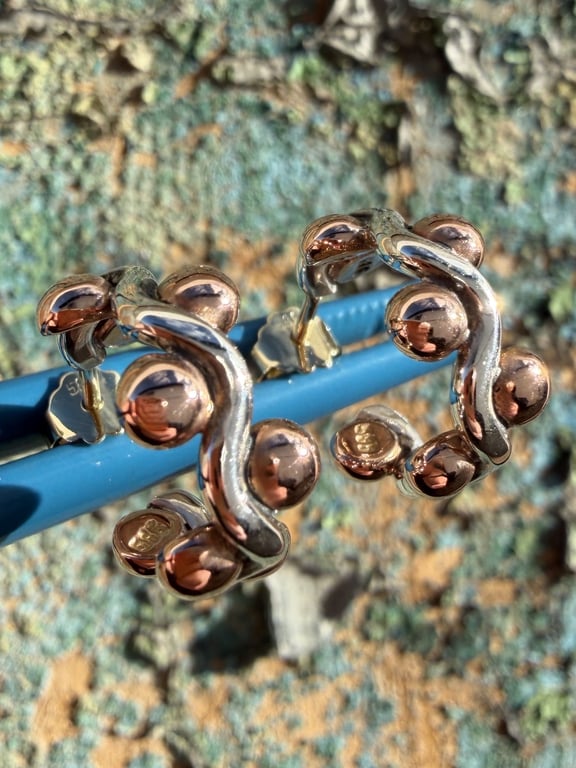 Nærbillede af unikke øreringe med bølget sølvdesign og runde kobberfarvede kugler, sat mod en blå baggrund.