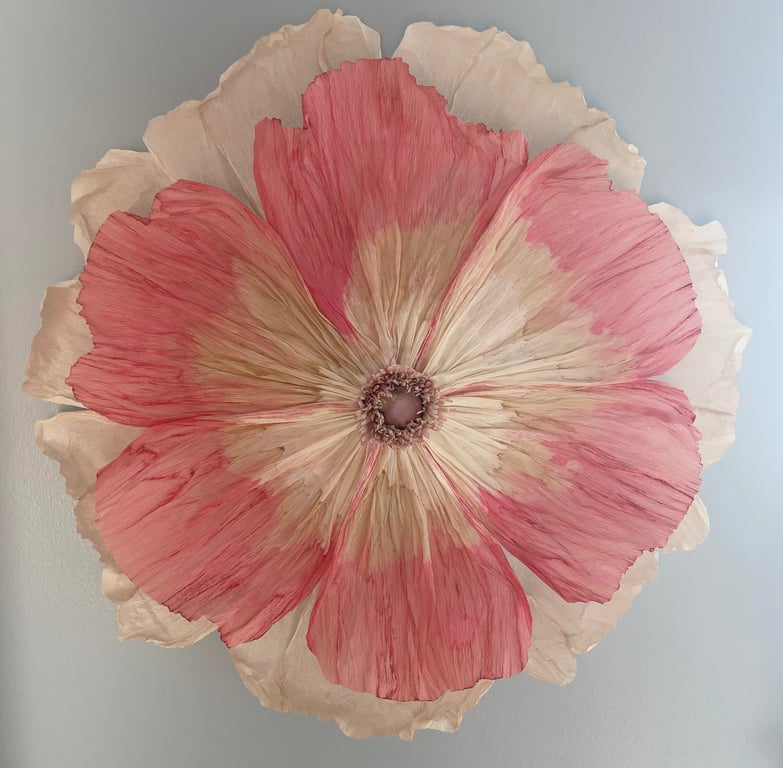 Stor, håndlavet blomst lavet af papir med lyserøde og beige kronblade på en lys væg.