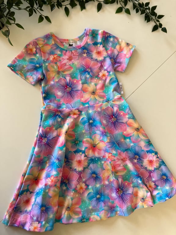 Farverig børnekjole med korte ærmer udsmykket med et mønster af store blomster i blå, lyserøde, lilla og gule nuancer, lagt ud på en lys overflade med grønt bladpynt øverst.