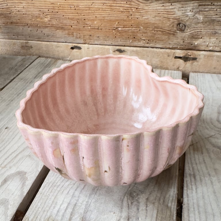 Pink keramisk skål formet som et hjerte med riller, placeret på et lyst træbord med en rustik trævæg i baggrunden.