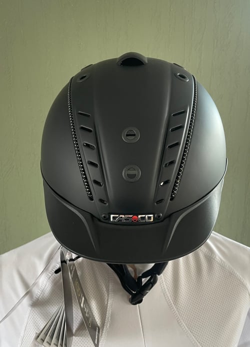 Sort ridehjelm med ventilationshuller og mærket "Casco" foran, båret af en person i hvidt tøj. (1)