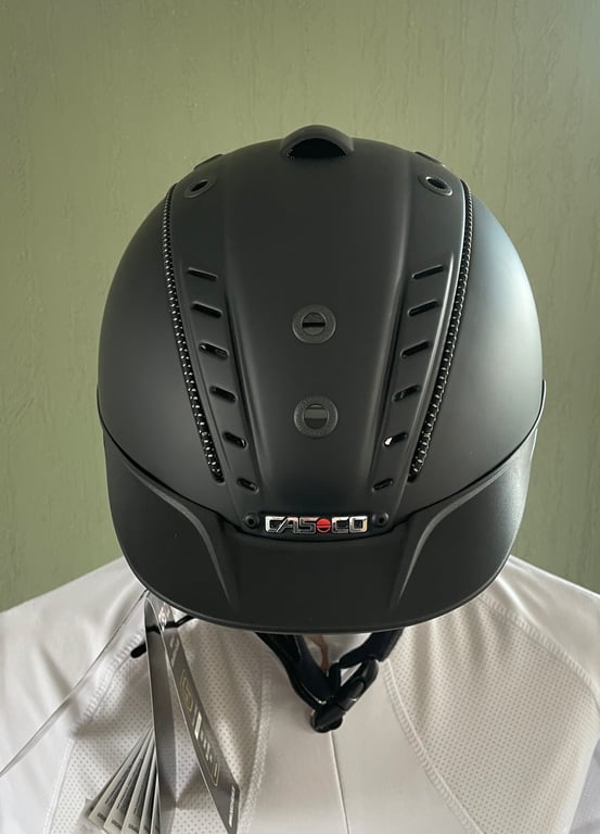 Sort ridehjelm med ventilationshuller og mærket "Casco" foran, båret af en person i hvidt tøj.