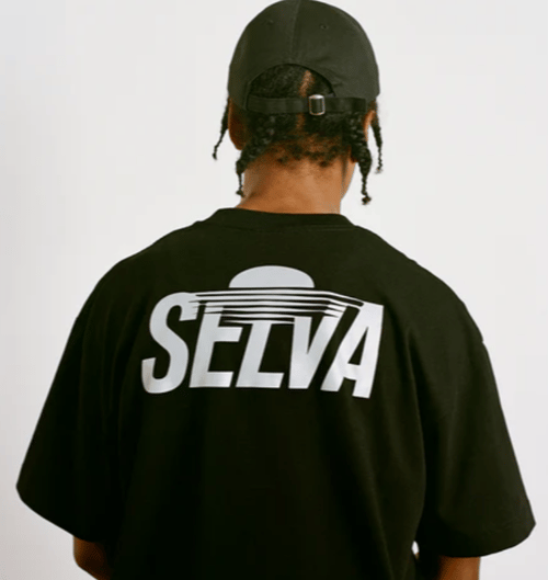 Mand med fletninger iført sort kasket og sort t-shirt med teksten "SELVA" på ryggen. (2)