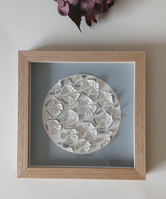 Indrammet papirklip med flere trekløves blade arrangeret i en cirkel på en blå baggrund, placeret på et hvidt bord med en lilla blomst øverst.