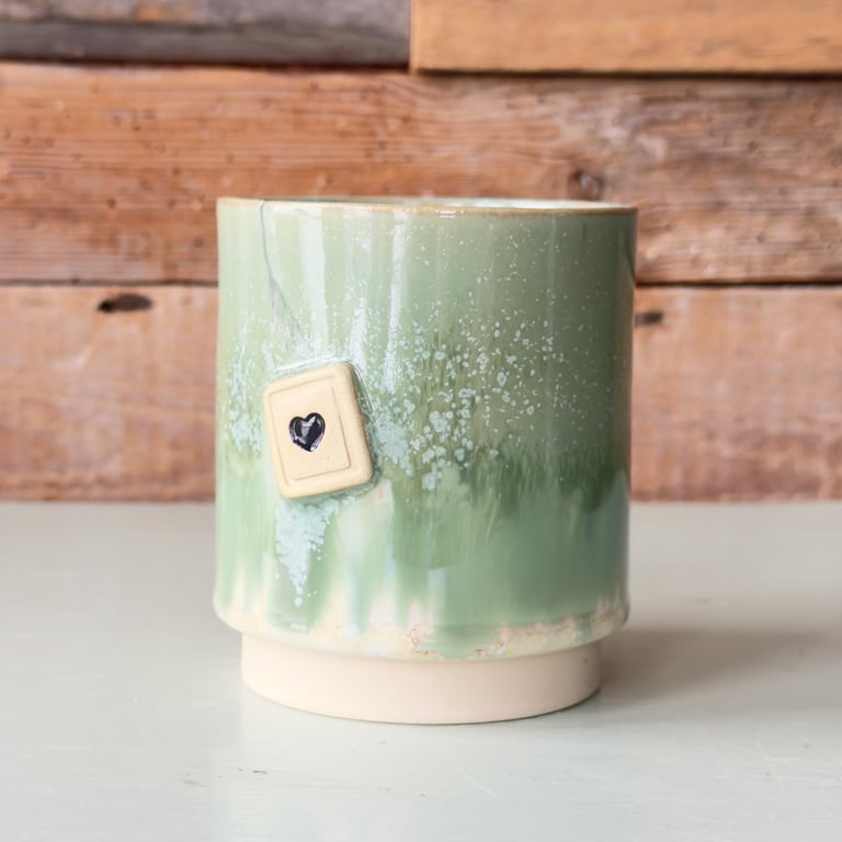 Grøn keramikkop med en lille firkantet mærkat med et sort hjerte, stående på en hvid overflade foran en trævæg.