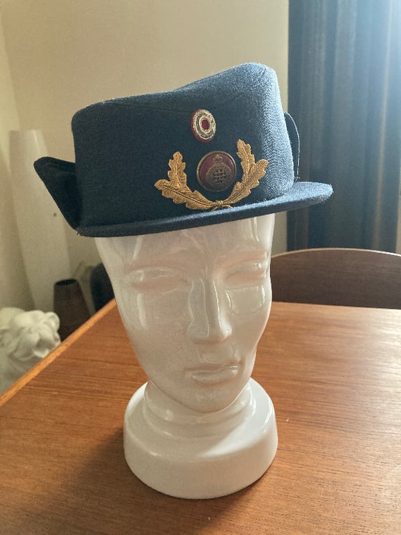 Billedet viser en mørkeblå militærhat med emblem, placeret på en hvid keramisk mannequinhoved på et træbord. (2)