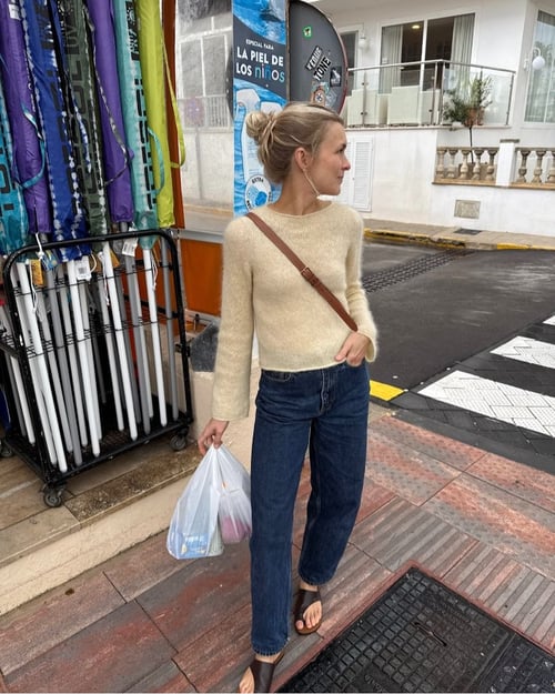 Kvinde iført beige striktrøje, blå jeans og brune sandaler, der bærer en plastikpose på en fortov ved en gade med farverige paraplyer til salg i baggrunden. (1)