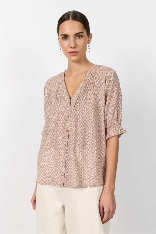 Kvinde i lyserød mønstret bluse med V-hals og korte ærmer samt lyse bukser mod hvid baggrund.