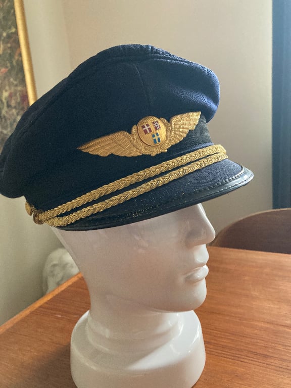 En mørkeblå pilotkasket med gyldne vinger og emblem samt to gyldne flettede bånd, placeret på en hvid mannekinehoved på et træbord. (2)