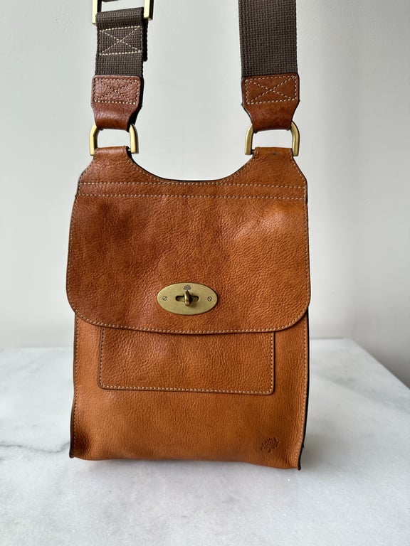 Brun lædertaske med flap og metalspænde, samt bred brun skulderrem med lærredsdetaljer.
