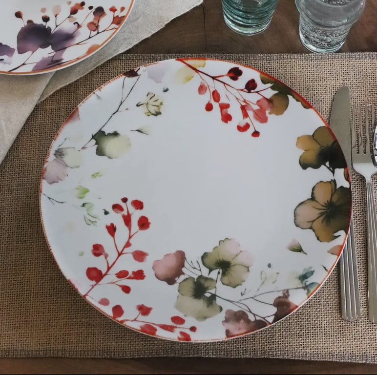 Rund flad tallerken med blomsterdekoration i røde, grønne og gule nuancer, placeret på en beige bordskåner sammen med bestik og glas. (2)