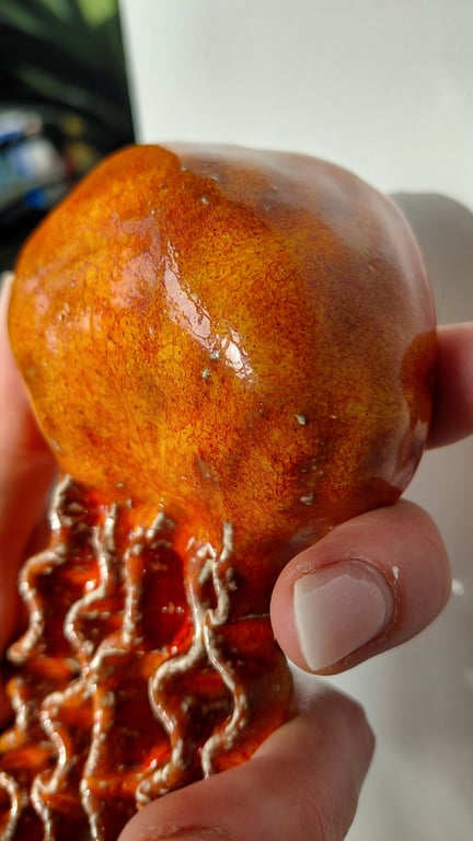 Hånd der holder en skinnende orange-rød keramisk figur med bølget mønster nederst.