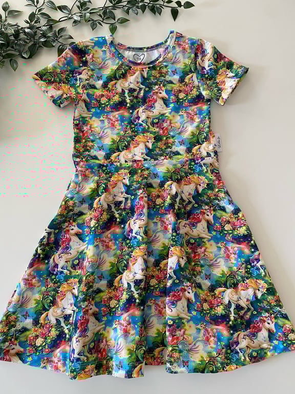 Børnekjole med korte ærmer og farverigt mønster af enhjørninger, blomster og regnbuer på blå baggrund, lagt fladt på hvid overflade med grønne blade ved siden af.