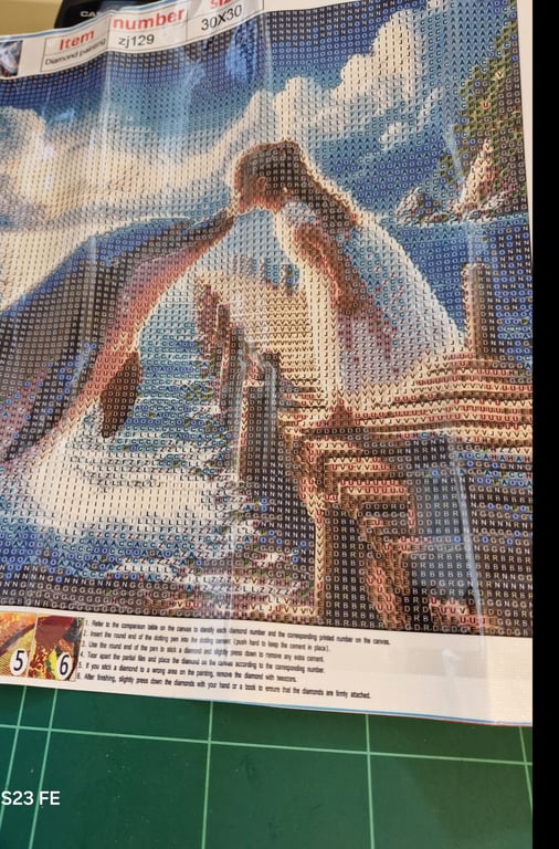 Et diamantmaleri under arbejdet, der forestiller en pige, der kysser en delfin ved havet med en træbro og klipper i baggrunden.