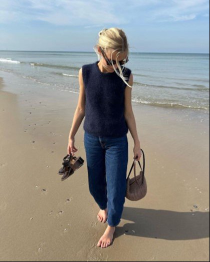 Kvinde med solbriller, mørk bluse og jeans går barfodet langs stranden, holder sko i den ene hånd og taske i den anden. (1)