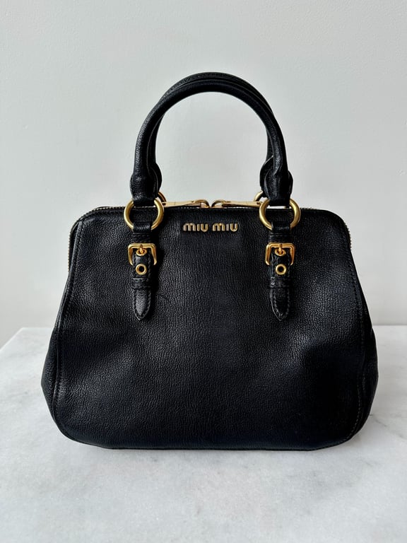 Sort Miu Miu håndtaske i læder med gyldne metaldetaljer og to håndtag, placeret på en hvid overflade.